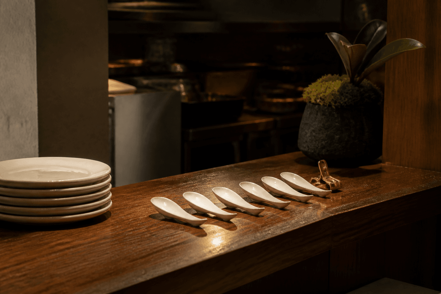 Unused tasting spoons in a row on a koa kitchen pass, unused ivory plates, brass clip.