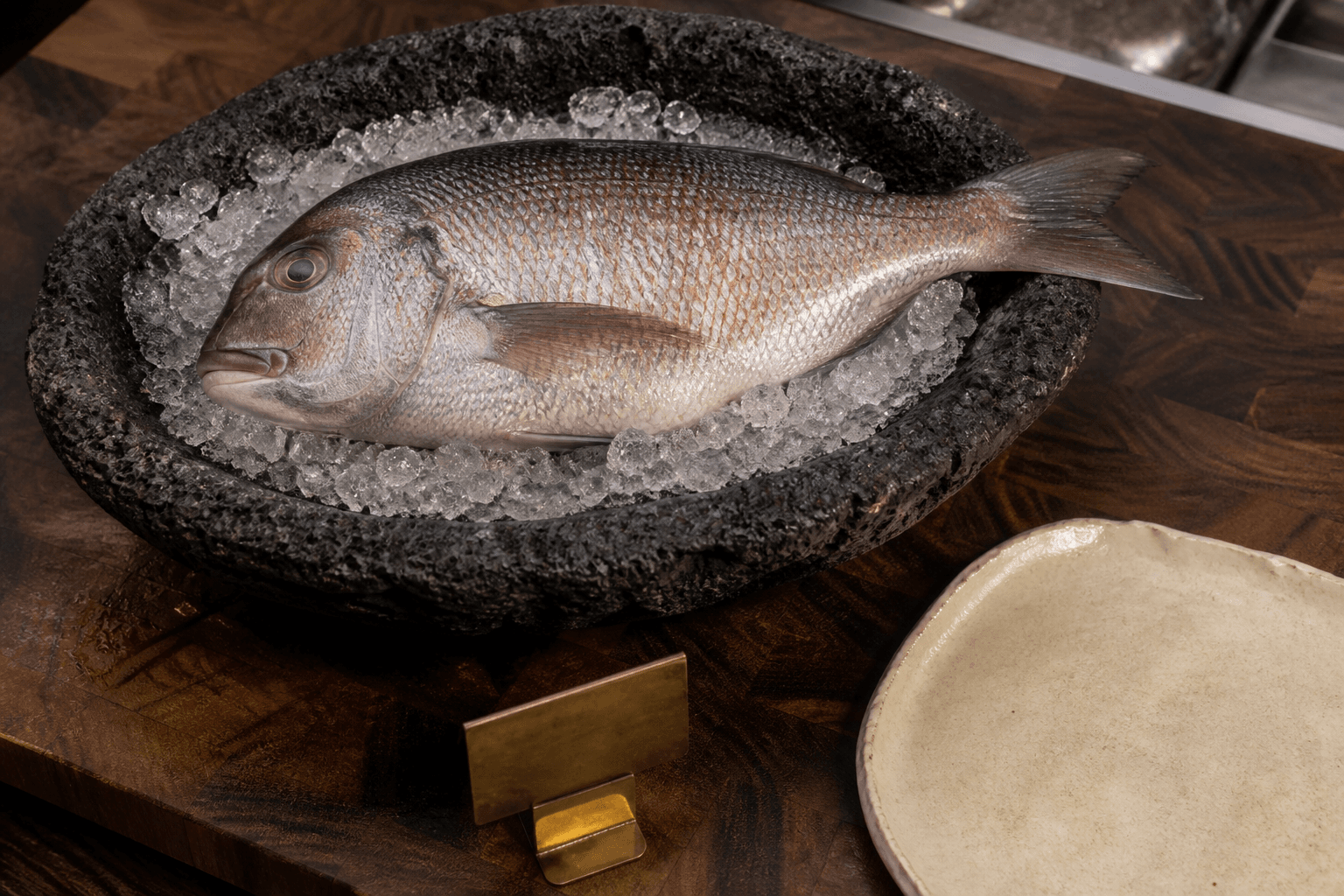 A whole unmarked fish on crushed ice in a volcanic-stone bowl, unused ivory plate, koa counter.