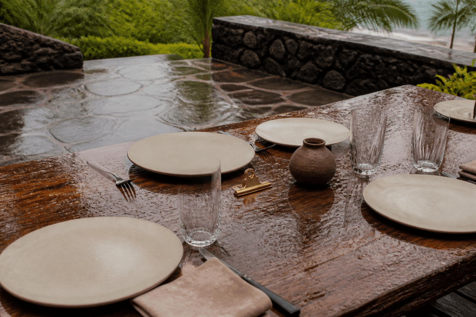 Unused ivory plates on a rain-dark stone terrace, wet koa table, empty glasses, brass clip.