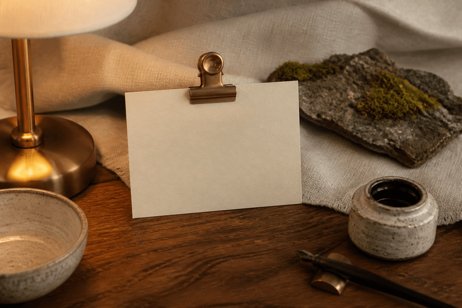 A blank ivory card in a brass clip on a koa desk, fountain pen, mossed lava, no writing.