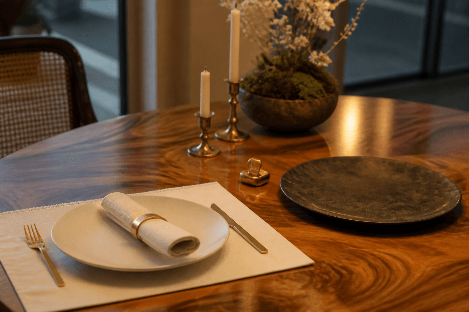 One unused ivory place setting beside an unused charger plate on a koa table, two unlit candles.